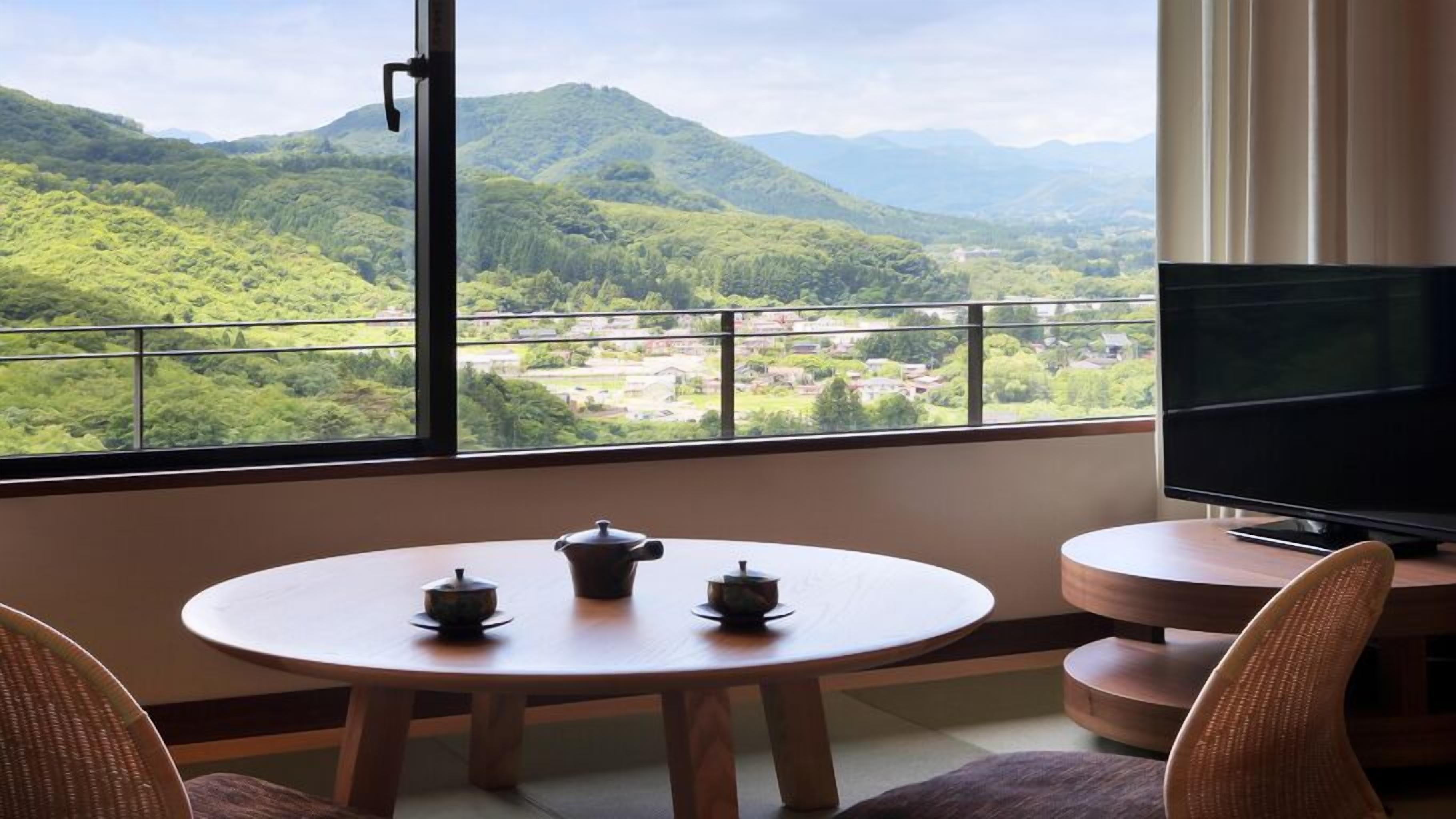 A room with a round table, two chairs, a TV, and a balcony with a view of green hills.