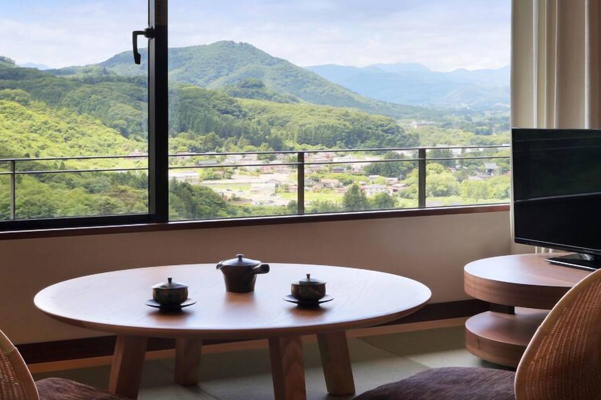 Una habitación con una mesa redonda, dos sillas, un televisor y un balcón con vistas a colinas verdes.