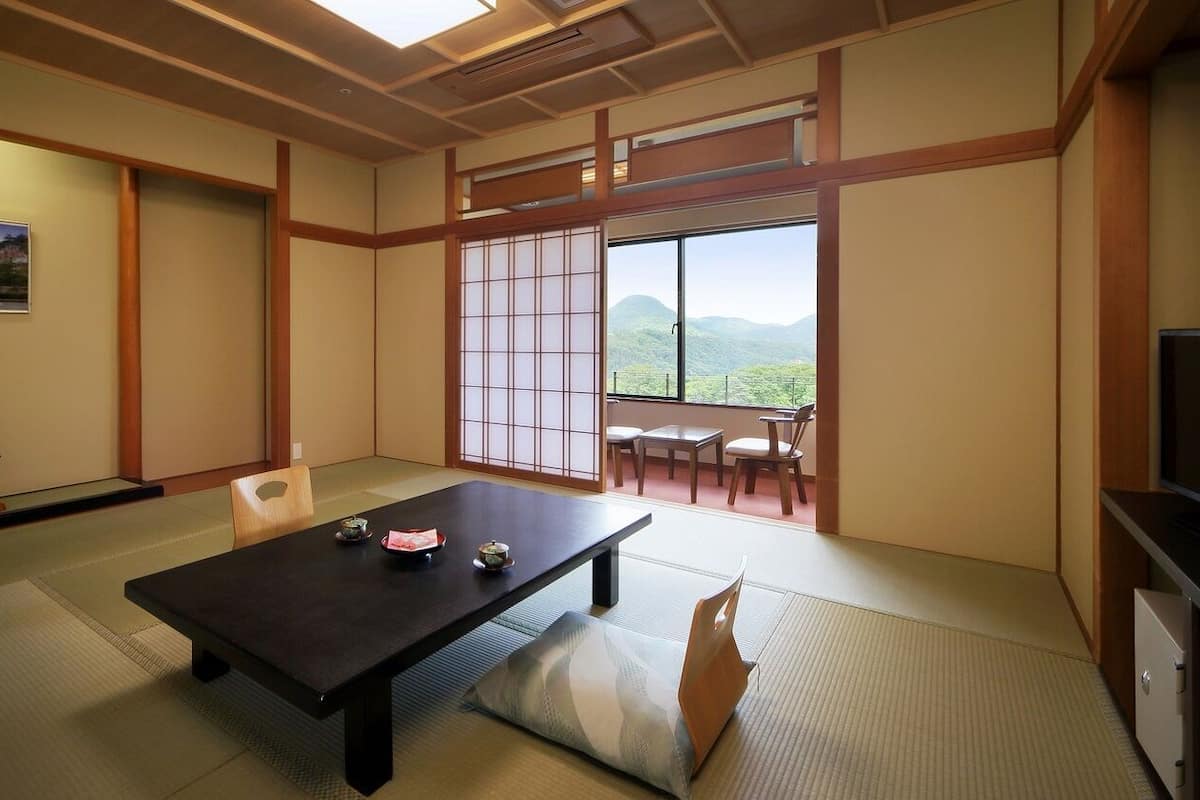 Una habitación tradicional japonesa con suelo de tatami, una mesa baja con cojín, una puerta corrediza con vistas y un televisor a la derecha.