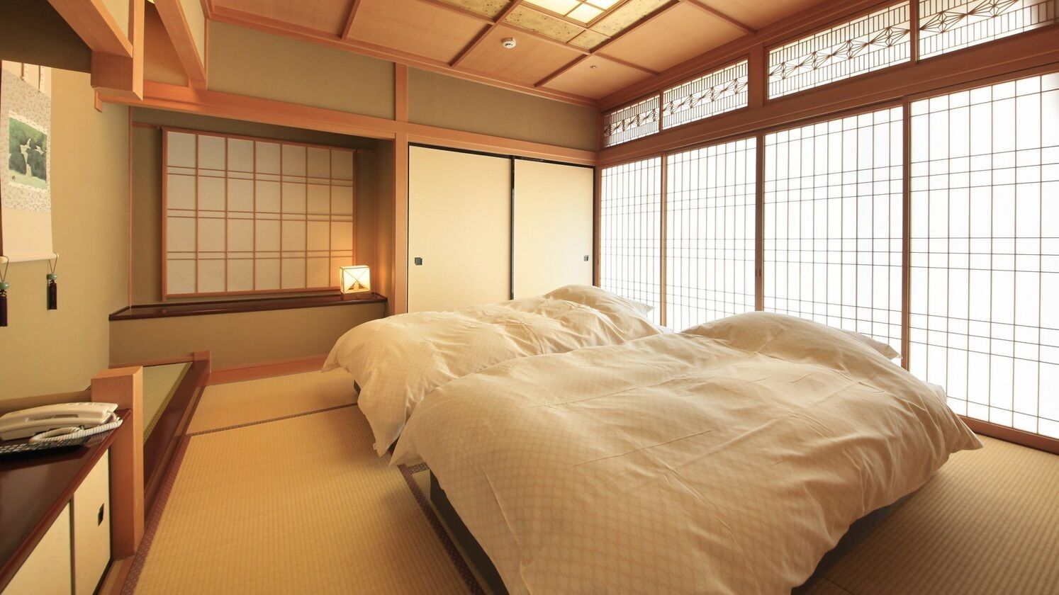 Una habitación de estilo japonés tradicional con suelo de tatami, una cama con ropa de cama blanca, puertas correderas y vistas a través de un celosía shoji.