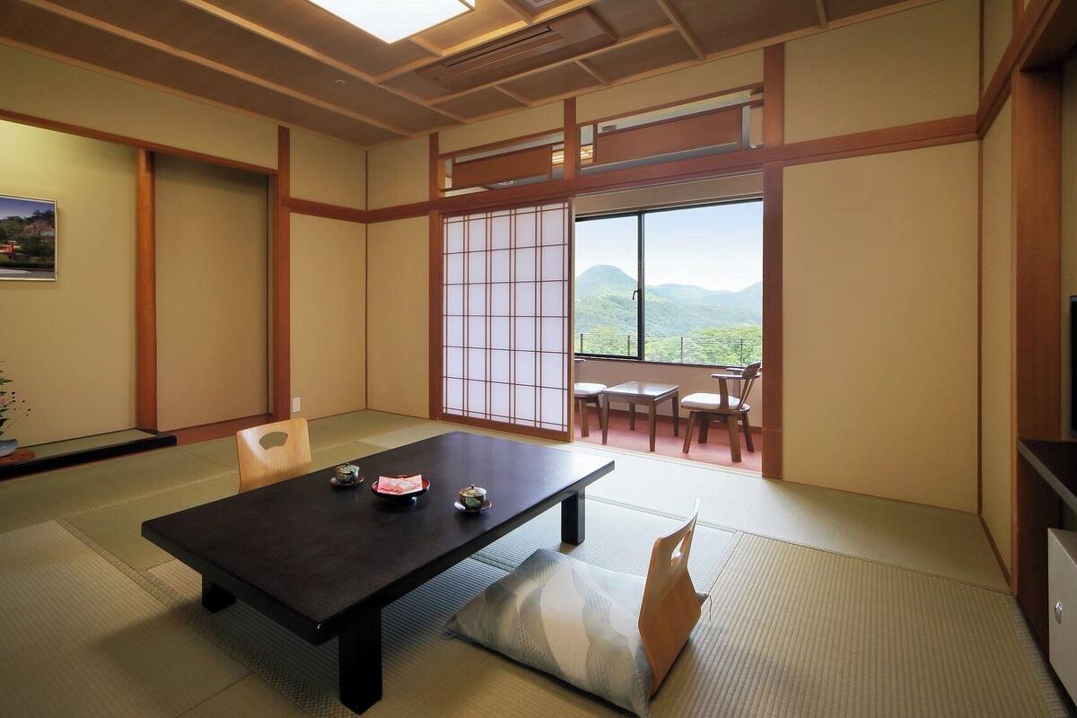 Una habitación japonesa tradicional con suelo de tatami, una mesa baja con cojines, puertas correderas y vistas a un paisaje montañoso.