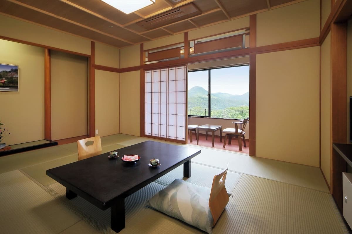 A traditional Japanese room with tatami flooring, a low table with cushions, sliding doors, and a view of a mountainous landscape.