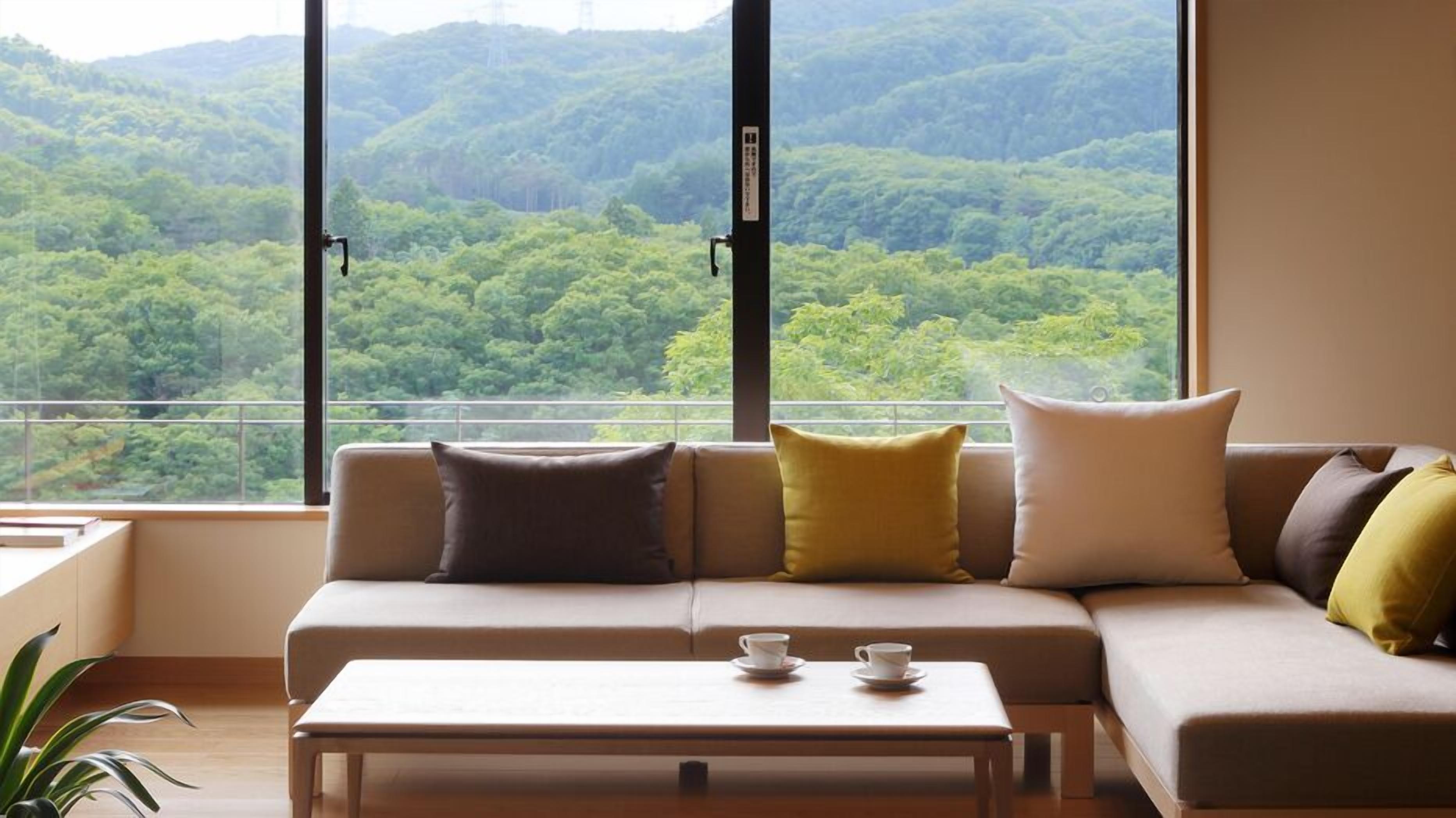 A modern living room with a sofa, cushions, a coffee table, and a view of a forested hillside.