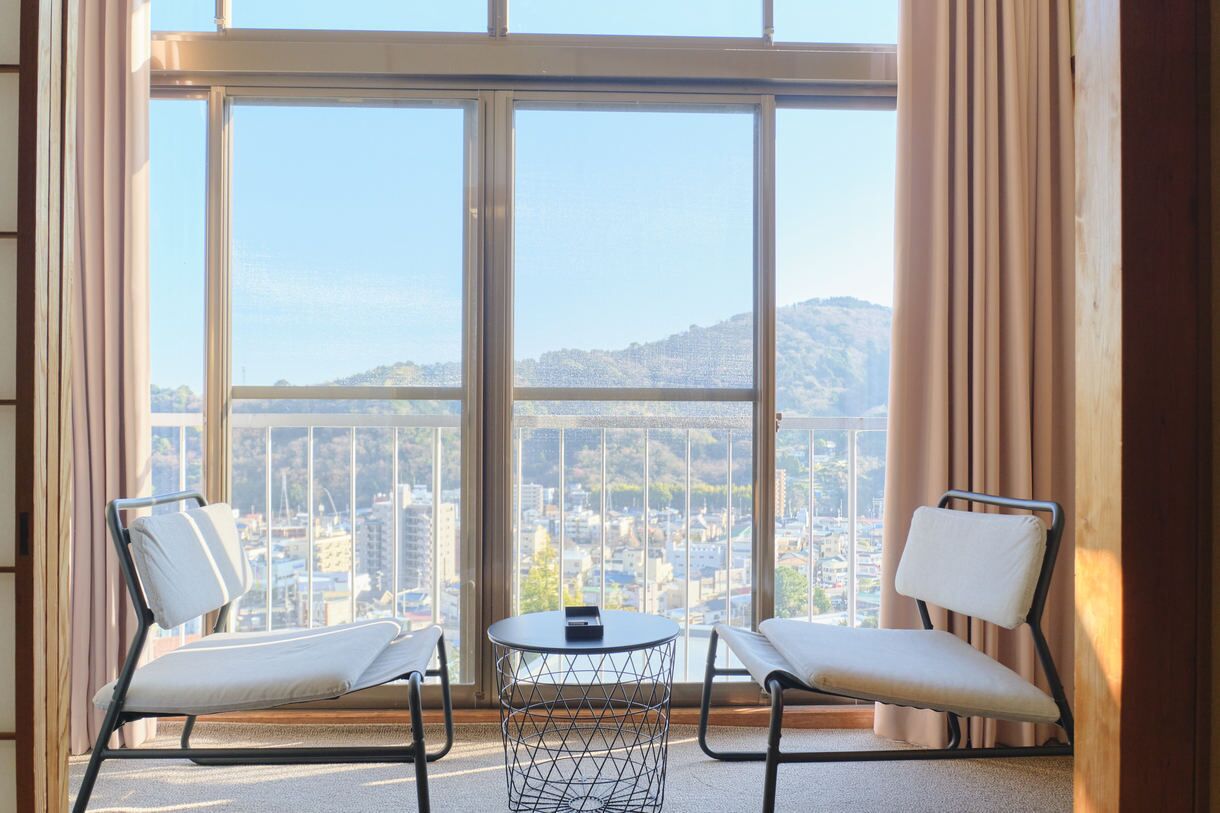 Un balcón con dos sillas y una mesita, con vistas a un paisaje urbano con edificios y colinas.