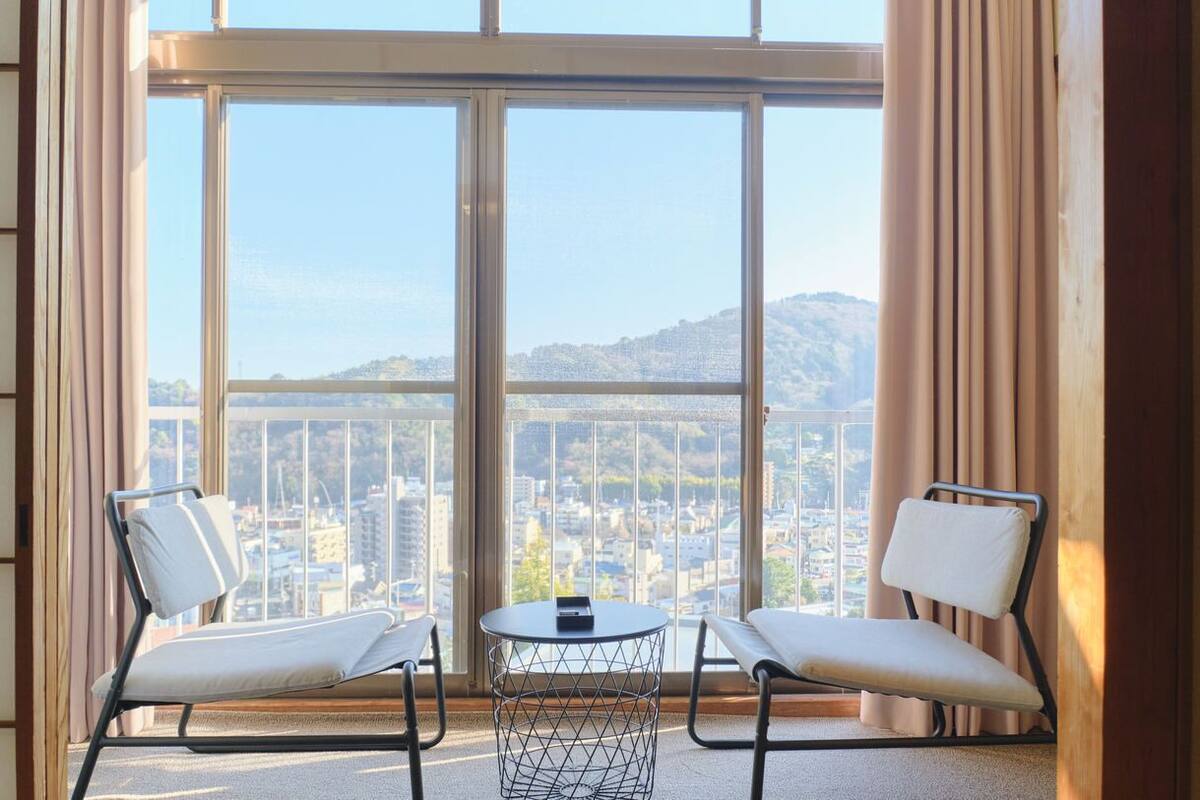 Un balcón con dos sillas y una mesita, con vistas a un paisaje urbano con edificios y colinas.