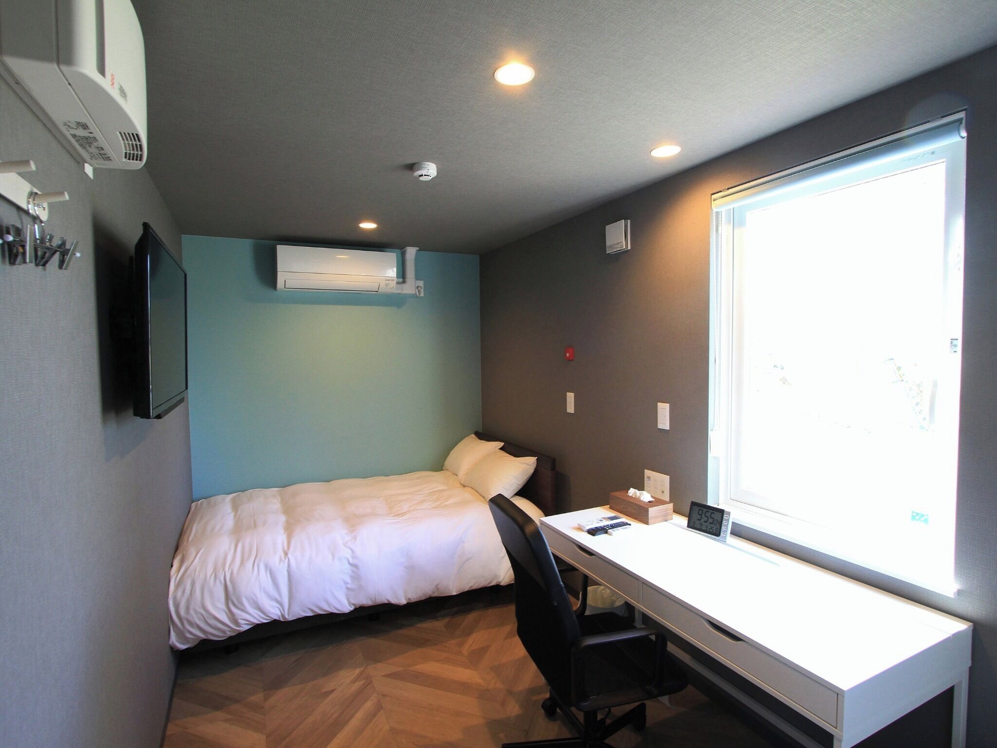 Una habitación pequeña y funcional con cama, escritorio y un ventanal amplio.