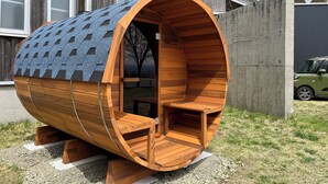A wooden barrel-shaped structure with a sloped roof, a bench inside, and a view of a house in the background. - Orando no Nikai (Aridagawa)
