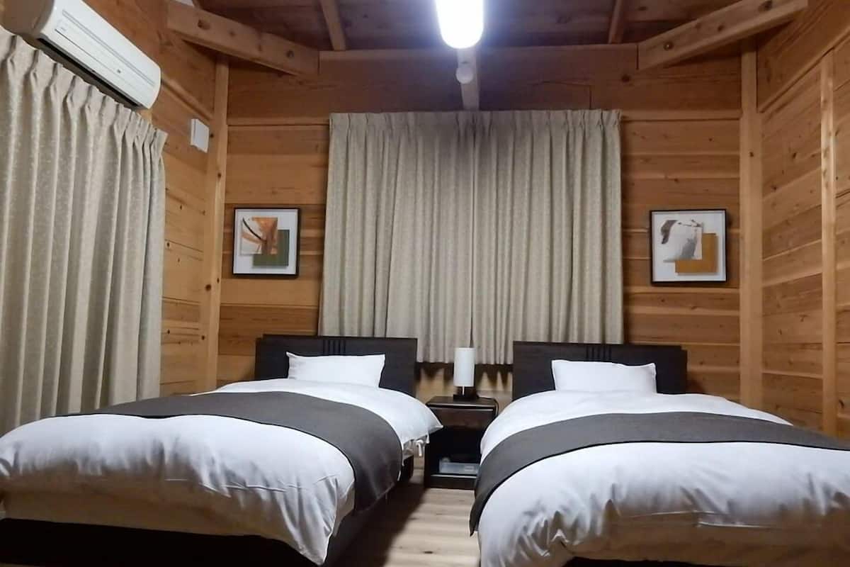 Ein Zimmer mit zwei Betten, Holzwänden, einem Deckenventilator und einem Fenster mit Vorhängen.