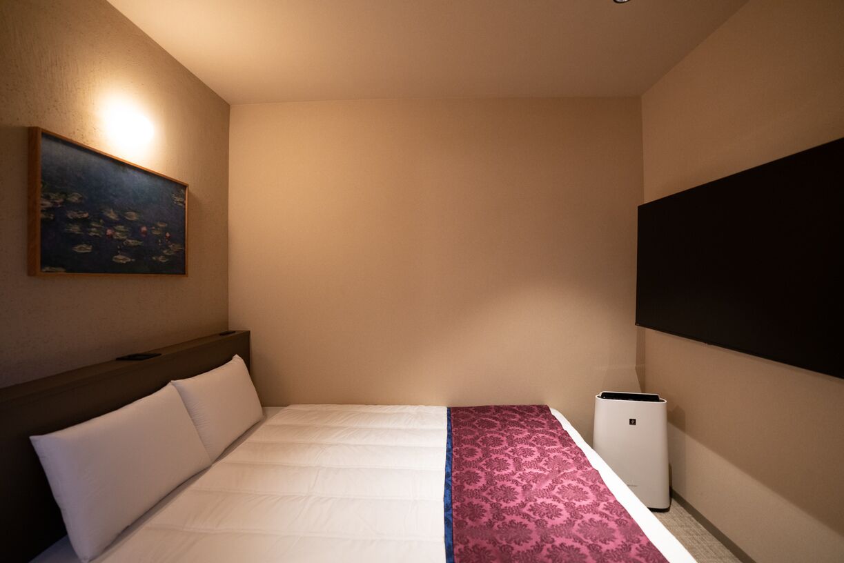 A hotel room with a bed, a painting on the wall, a large window, and a wall-mounted air conditioner.