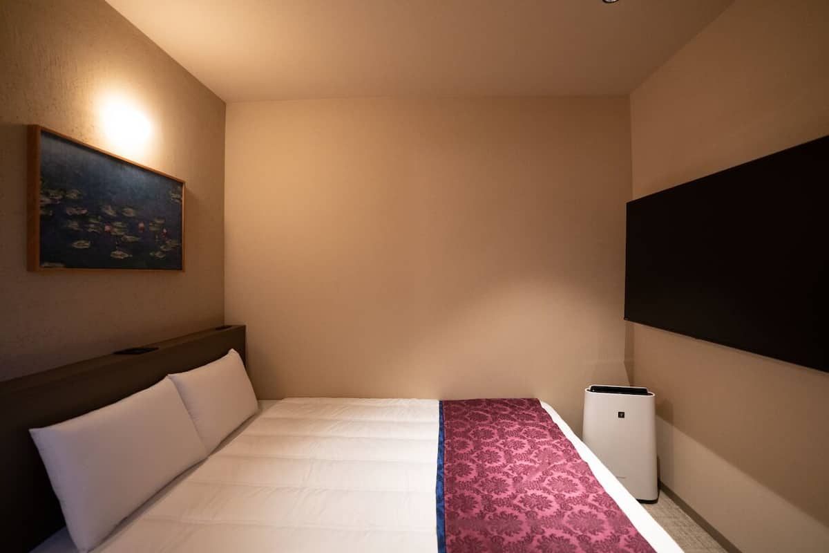 Ett hotellrum med en säng, en tavla på väggen, ett stort fönster och en väggmonterad luftkonditioneringsanläggning.