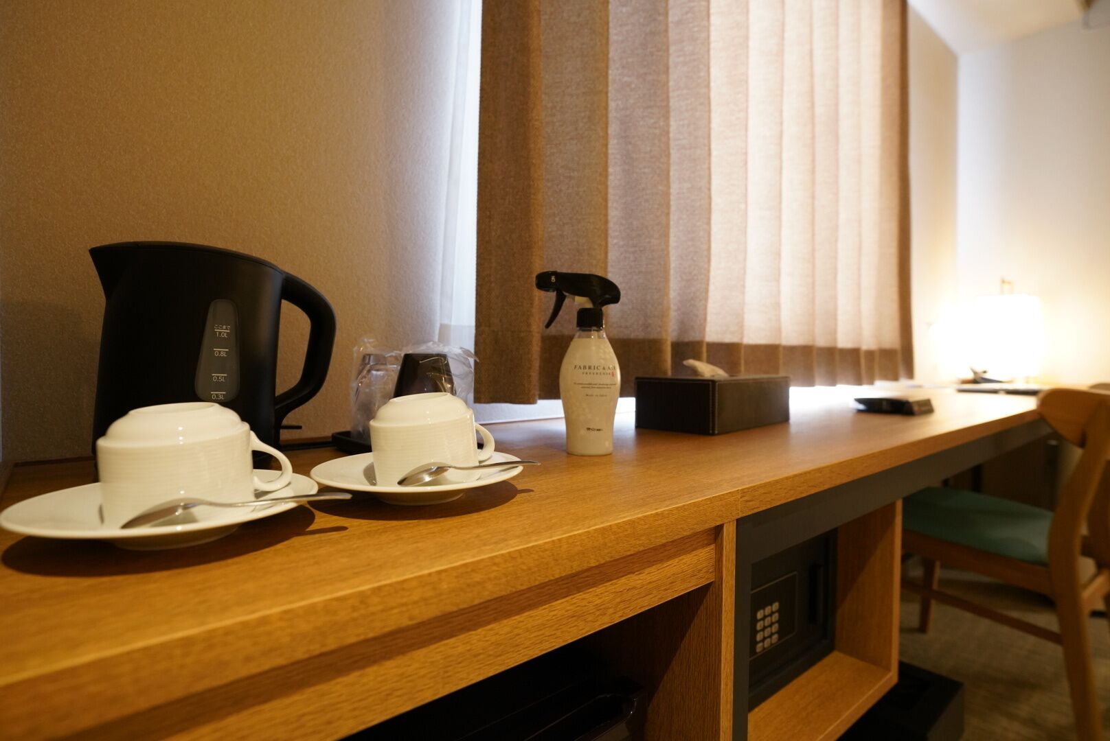 A hotel room with a wooden desk, two white cups, a coffee maker, a spray bottle, and a remote control.