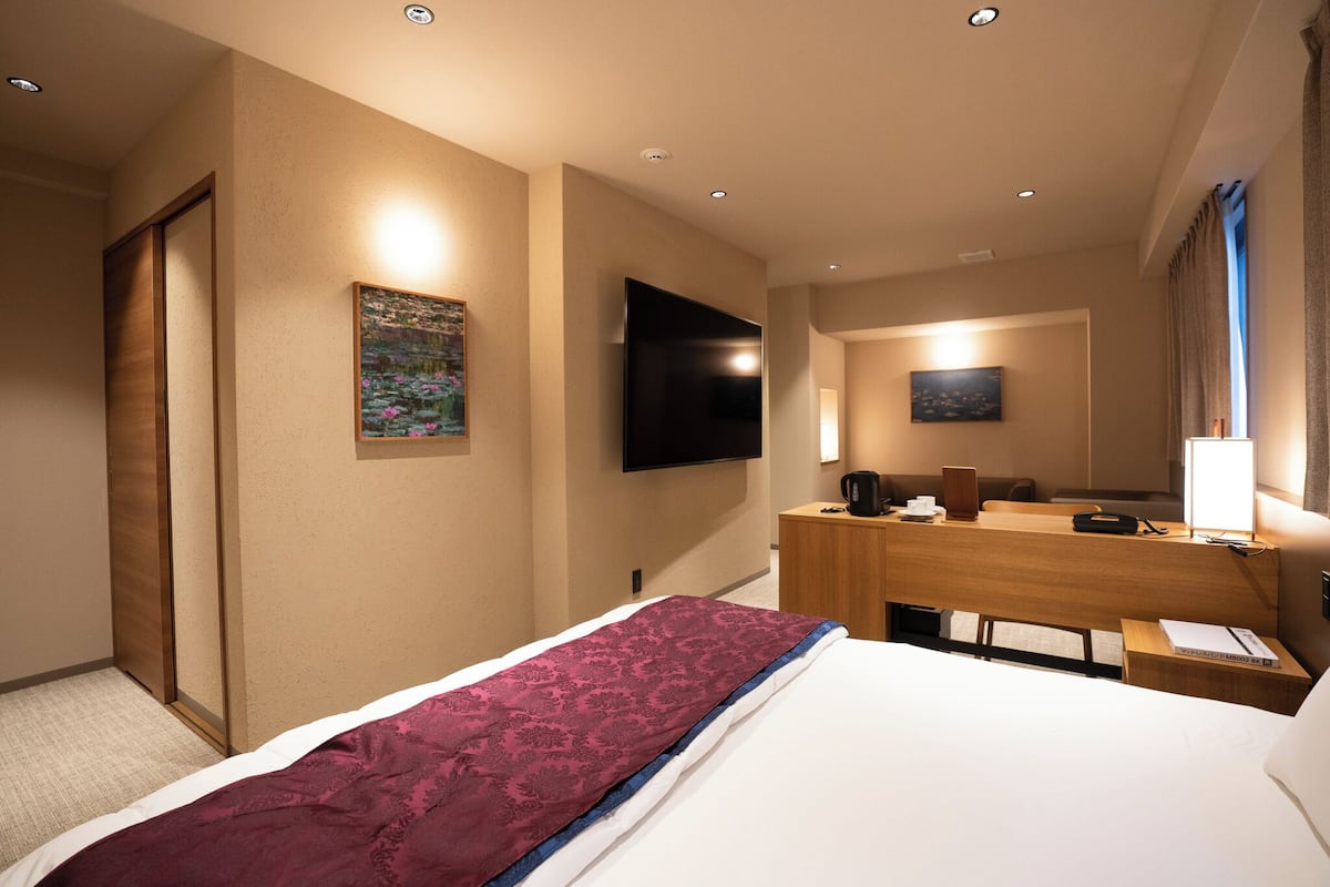 Ett hotellrum med en säng, ett skrivbord, en platt-TV, en tavla och ett fönster med gardiner.