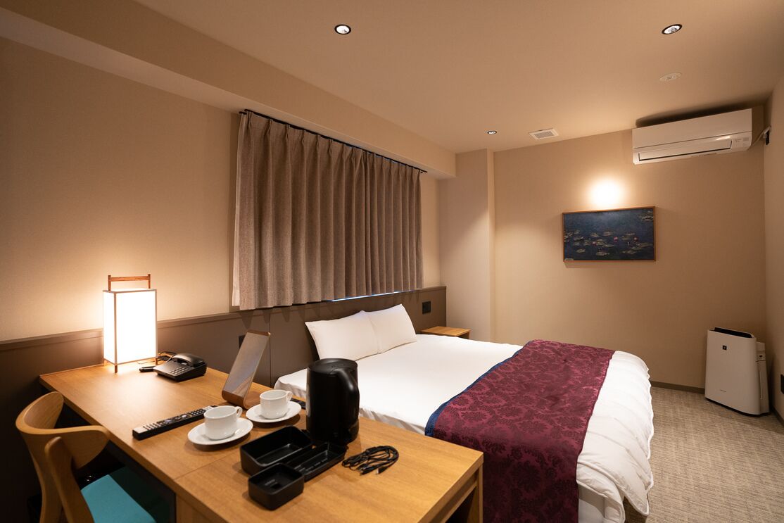 A hotel room with a bed, a desk with a lamp, a coffee maker, a remote control, a telephone, and a painting on the wall.