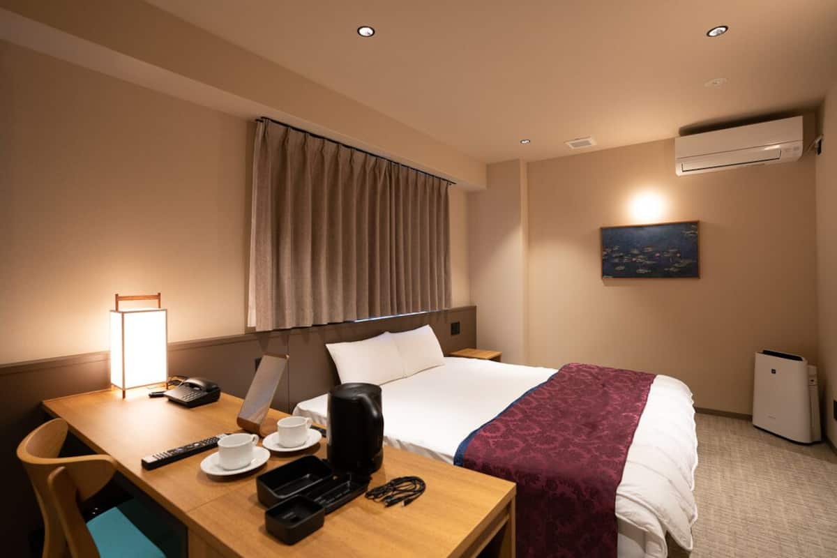 Ett hotellrum med en säng, ett skrivbord med en lampa, en kaffebryggare, en fjärrkontroll, en telefon och en tavla på väggen.
