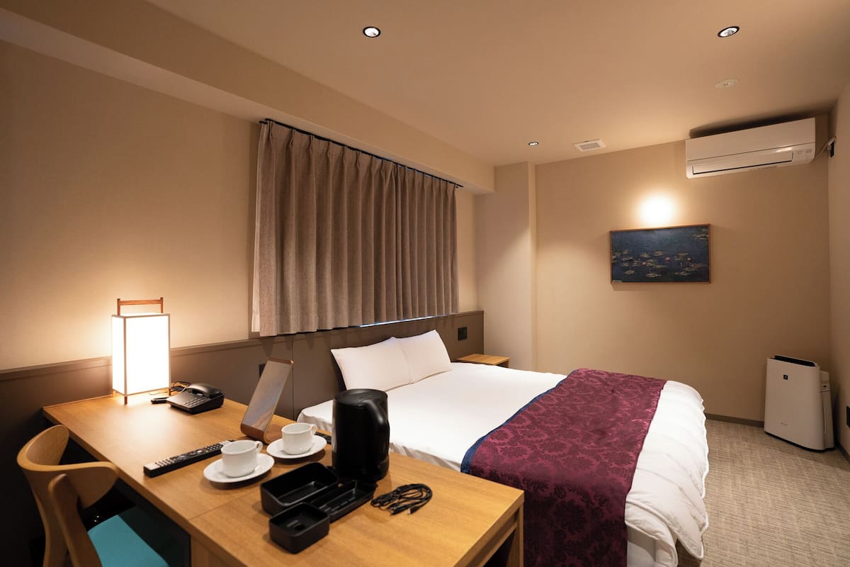 A hotel room with a bed, bedside table, lamp, air conditioning unit, and a painting on the wall.