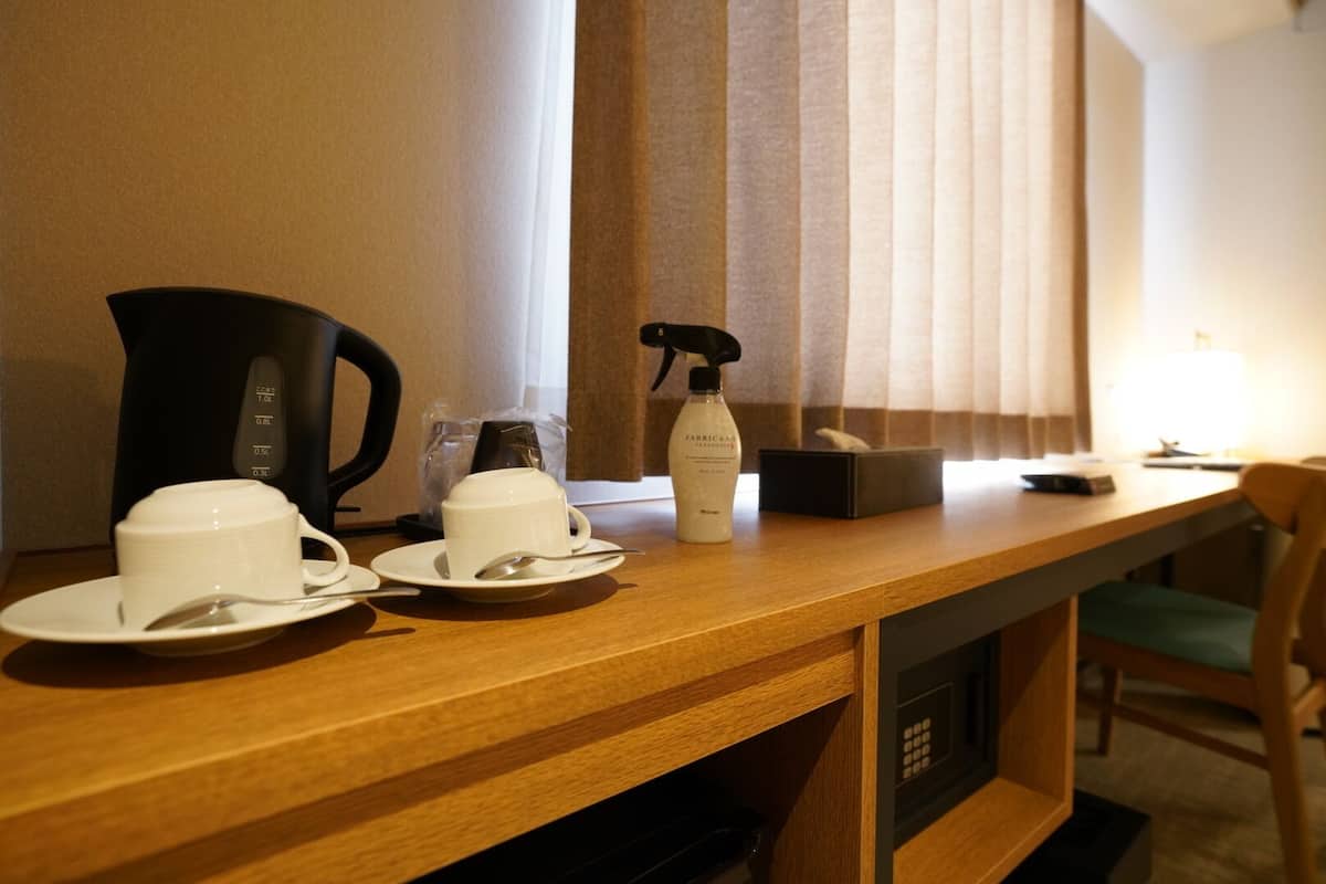 A hotel room with a wooden desk, two white cups on saucers, a coffee maker, a spray bottle, a remote control, and a window with blinds.