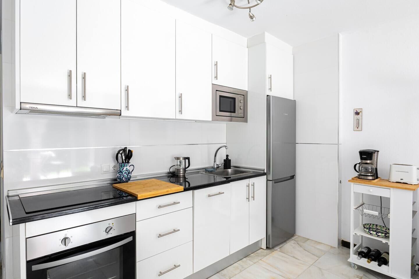 Een moderne keuken met witte kasten, een roestvrijstalen koelkast, een magnetron en een koffiezetapparaat op een klein houten tafeltje.