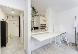 A modern kitchen with white cabinetry, a white countertop, and a stainless steel sink. There is a white stool and a white cabinet in the corner. - Azory by Park 2-Bedroom Apartment (Kraków)