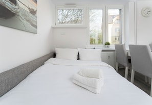 A neatly made bed with white linens, a grey headboard, and a window with a view of buildings and trees. - Azory by Park 2-Bedroom Apartment (Kraków)