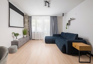 Ein modernes Wohnzimmer mit einem Ecksofa, einem Holztisch, einem grauen Ottomanen, einer Topfpflanze, einem Wand-Fernseher und einer Deckenleuchte.