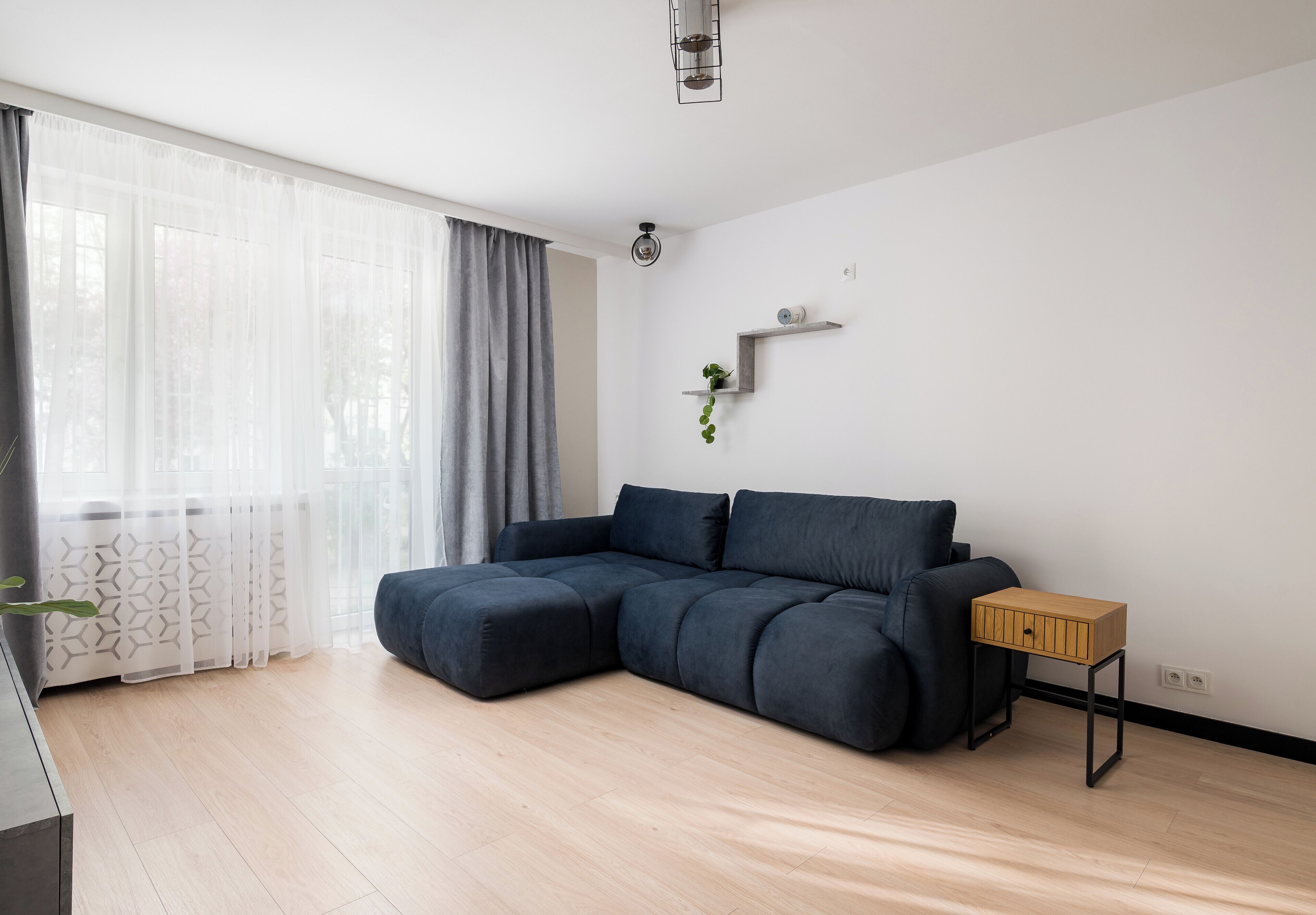 A modern living room with a sectional sofa, a small wooden side table, a potted plant, and a window with sheer curtains.