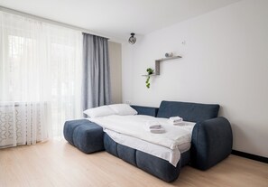 Ein modernes Zimmer mit Schlafsofa, einer kleinen Pflanze auf einem Regal und einem Fenster mit durchsichtigen Vorhängen.