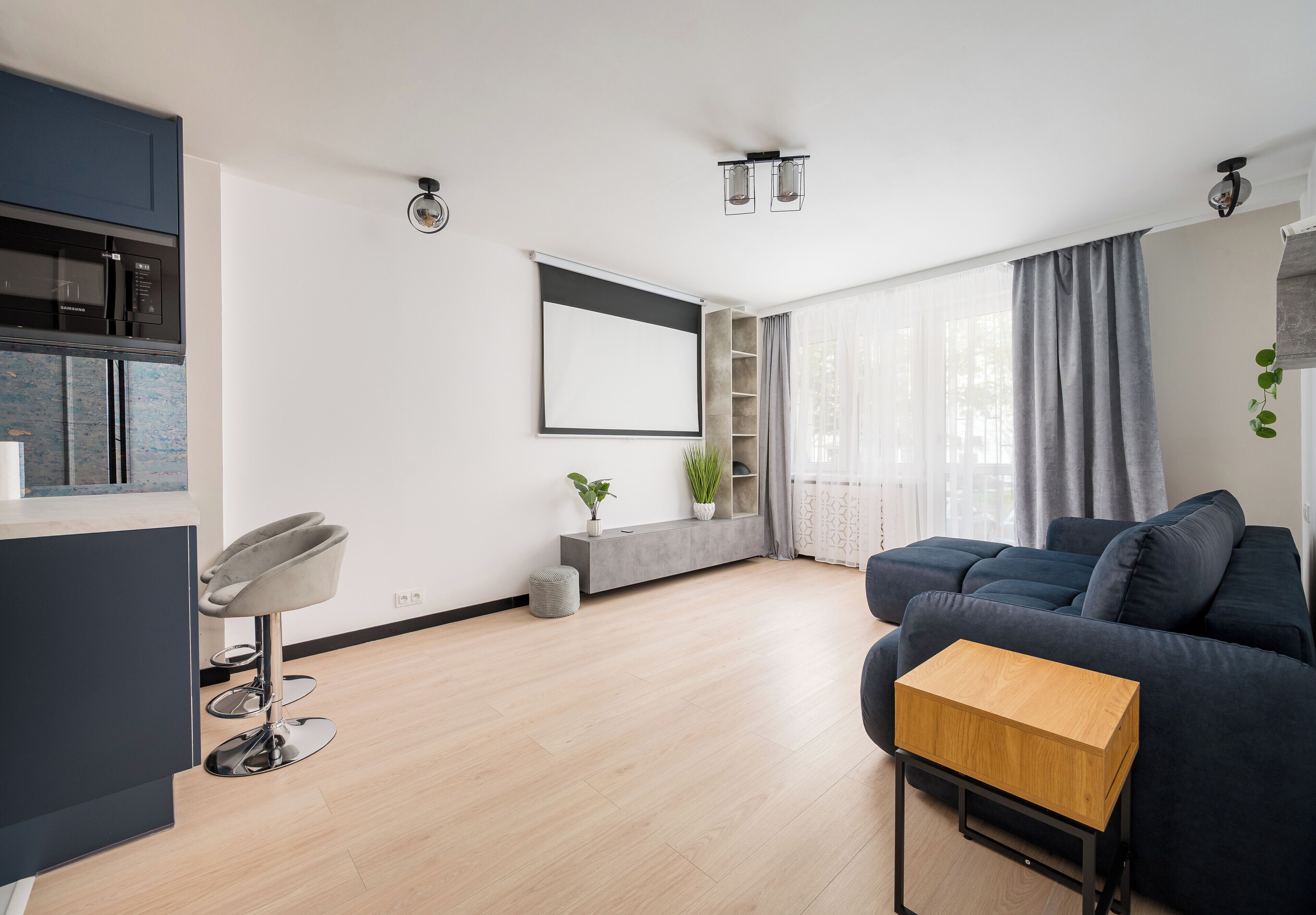 A modern living room with a dark sofa, a wooden coffee table, a kitchen area with a microwave, and a large window with curtains.