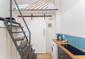 Ein kompaktes Zimmer mit Etagenbett, Waschmaschine, einer kleinen Küchenzeile und einer Holztreppe.