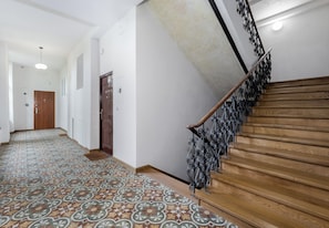 Ein Flur mit gemustertem Boden, einer Holztreppe mit Metallgeländern und einer Tür am Ende.