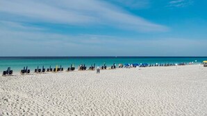 Een breed zandstrand met rijen ligbedden en parasols, een heldere blauwe lucht en een rustige zee.