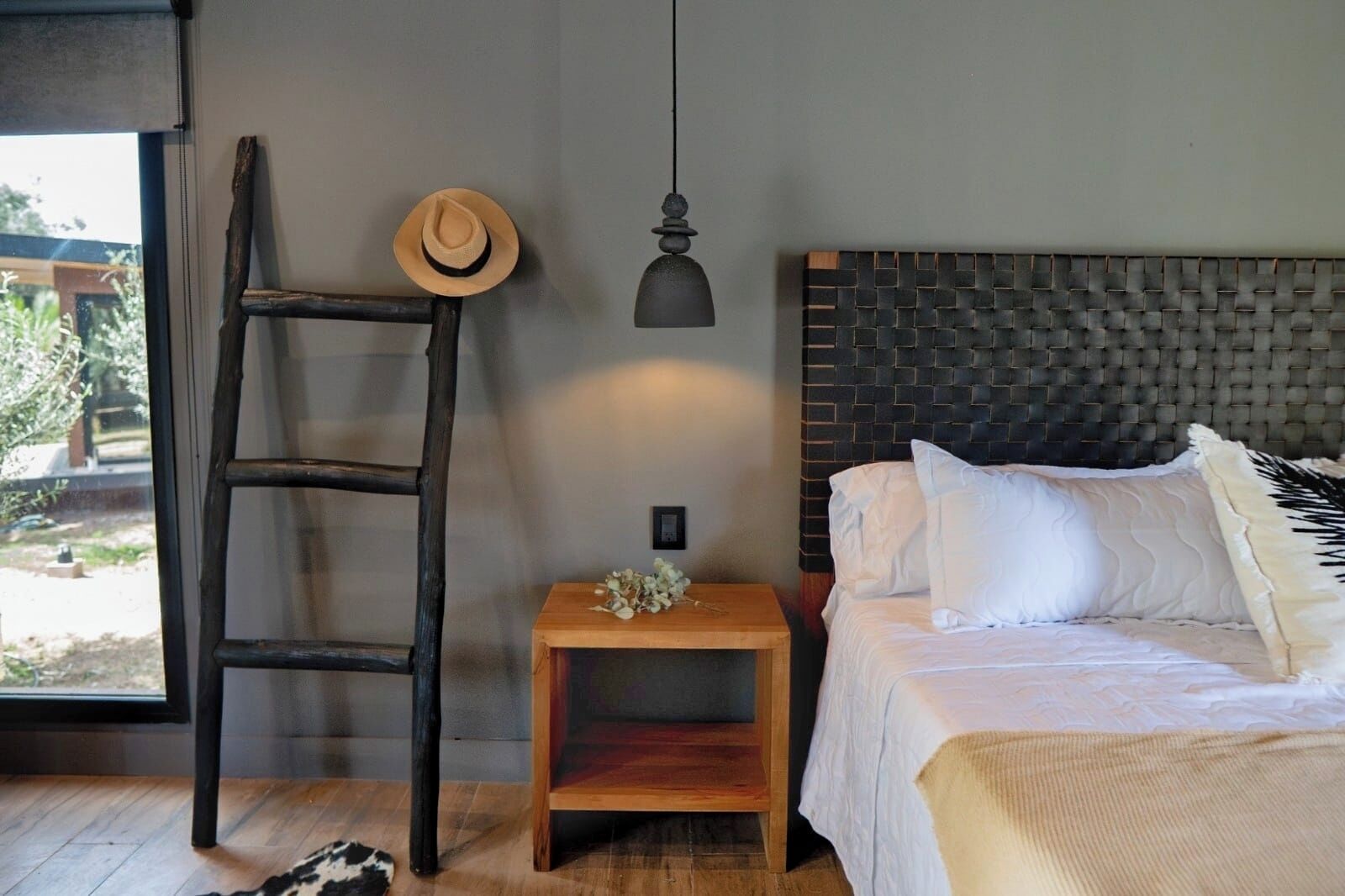 A bedroom with a wooden ladder, a bedside table with a hat, a hanging lamp, and a bed with white bedding.