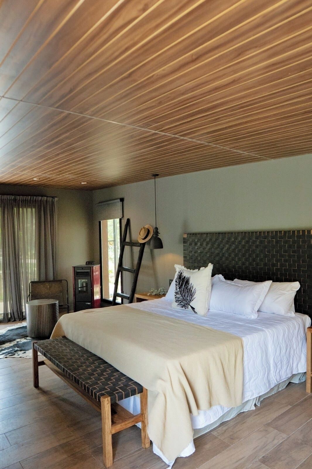 Een slaapkamer met een houten plafond, een bed met witte beddengoed, een bankje, een ladder en een bureau met een lamp.
