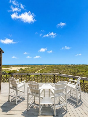 Eine Holzterrasse mit vier weißen Adirondack-Stühlen und einem Tisch, mit Blick auf Strand und Meer.