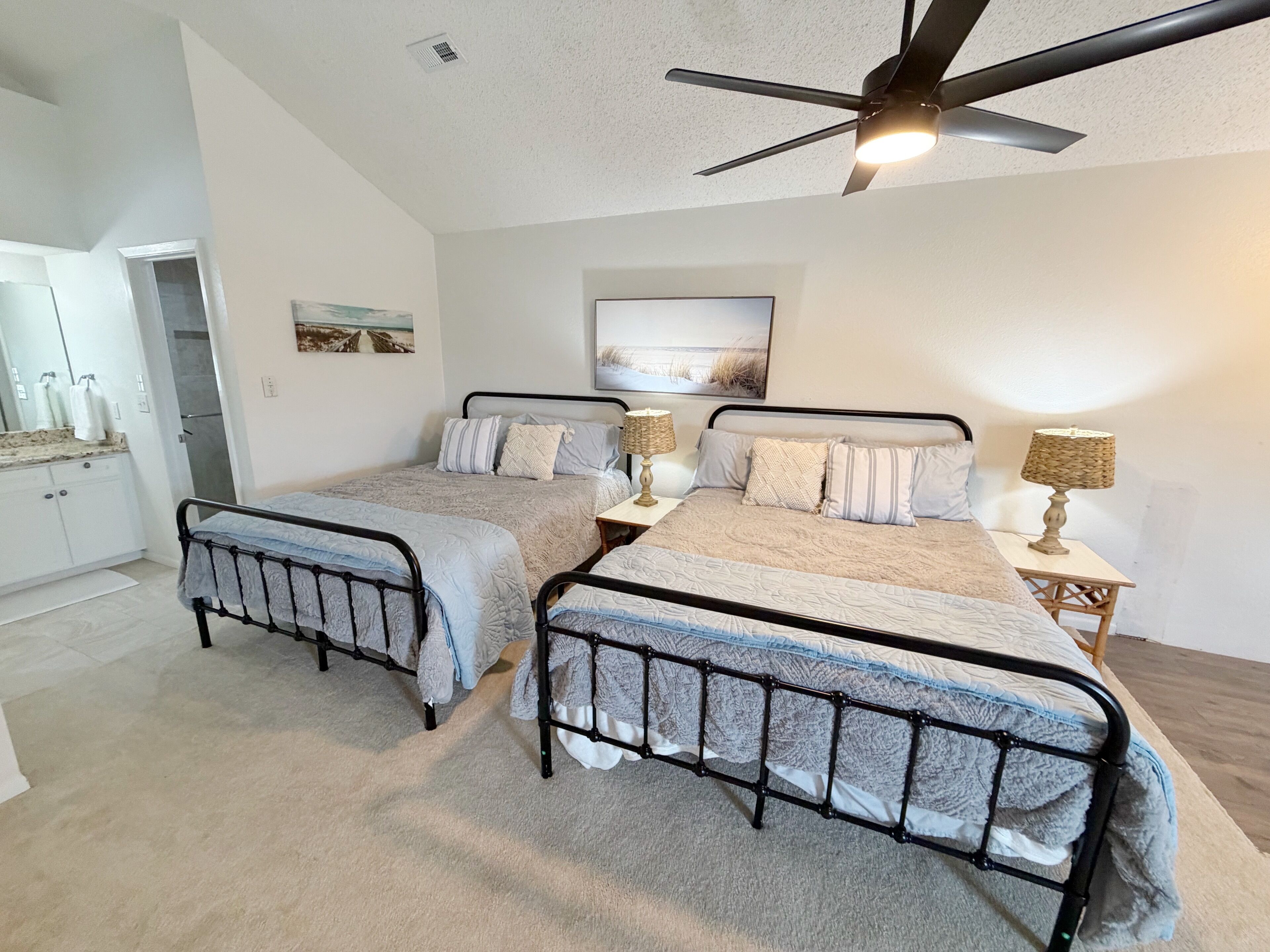 A bedroom with two beds, a ceiling fan, a painting, and a small table with a lamp.