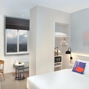 A modern hotel room with a large bed, a small table with a chair, a window with a city view, and a wardrobe with hanging space. - Fox Lite Hotel Samarinda (Samarinda)