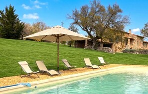 Ein Pool mit Liegestühlen und einem Sonnenschirm, umgeben von einer Rasenfläche und einem Steinhaus.