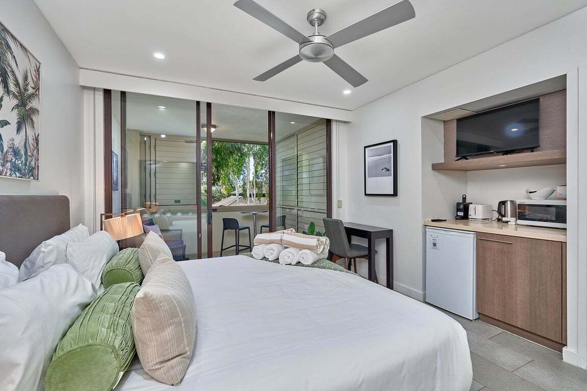 A modern hotel room with a bed, a desk, a television, and a kitchenette.