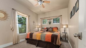 Ein Schlafzimmer mit einem Bett, zwei Nachttischlampen, einem Deckenventilator, einem Fenster mit Vorhängen und einem an der Wand befestigten Kruzifix.
