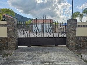 Ein verschlossenes Tor mit schmiedeeisernen Verzierungen, Steinpfeilern und Blick auf einen Berg sowie ein Gebäude mit rotem Ziegeldach.