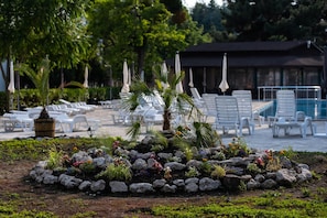 Zona junto a la piscina con sillones blancos y sombrillas, rodeada por un jardín de rocas con diversas plantas.