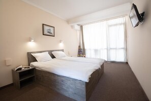 Een hotelkamer met een tweepersoonsbed, nachtkastjes, een wandtelevisie en een groot raam met gordijnen.