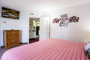 Ein Schlafzimmer mit einer rosa gesteppten Bettdecke, einem Holzschrank, einem an der Wand montierten Fernseher und einem Schild mit der Aufschrift 'GONE HIKING' und gerahmten Fotos.