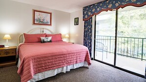 A bedroom with a bed, a nightstand, a lamp, a painting, and a balcony with a chair. - King 1BR Retreat, Jetted Bathtub, Indoor + Outdoor Pool, Walk to Parkway (Gatlinburg)