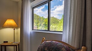 A room with a bed, a lamp, a table with a clock, and a window with a view of a green hill. - King 1BR Retreat, Jetted Bathtub, Indoor + Outdoor Pool, Walk to Parkway (Gatlinburg)