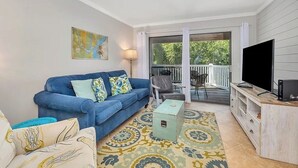 A living room with a blue sofa, a coffee table, a TV, and a balcony with outdoor furniture. - Coastal Condo with Partial Ocean View | Walk to East Beach | Pool + Grills (Saint Simons Island)