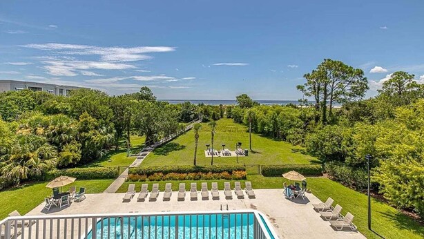 A pool area with lounge chairs, a well-manicured lawn, and a view of the ocean. - Coastal Condo with Partial Ocean View | Walk to East Beach | Pool + Grills (Saint Simons Island)