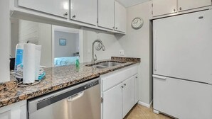 A kitchen with white cabinets, a stainless steel dishwasher, a granite countertop, a sink, and a clock on the wall. - Coastal Condo with Partial Ocean View | Walk to East Beach | Pool + Grills (Saint Simons Island)