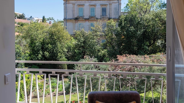 A view from a balcony with a railing, overlooking a large building with multiple chimneys and windows, surrounded by lush greenery. - Bright 2-Bedroom Apartment – Close to La Croisette & City Centre (Cannes)