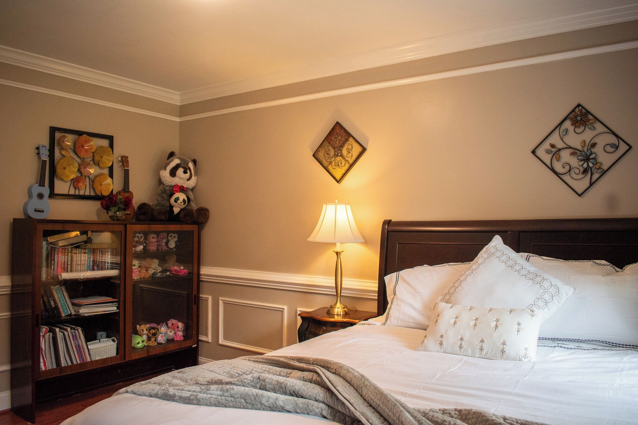 Een netjes opgemaakt bed met witte lakens en decoratieve kussens, een houten boekenplank vol boeken en speelgoed, een wandlamp en een lijst met een afbeelding boven het bed.