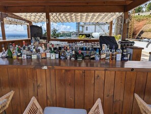 Un bar avec un comptoir en bois, diverses bouteilles et une vue sur l’océan et les bâtiments.